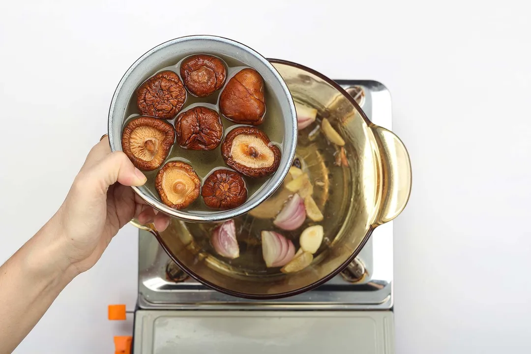 A bowl containing shiitake mushrooms and water being poured into a saucepan