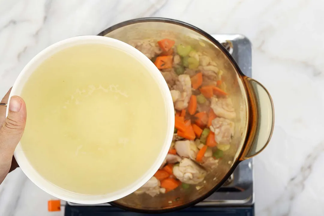 hand holds a bowl of chicken broth on top of a pot of cooked chicken and carrot cubes