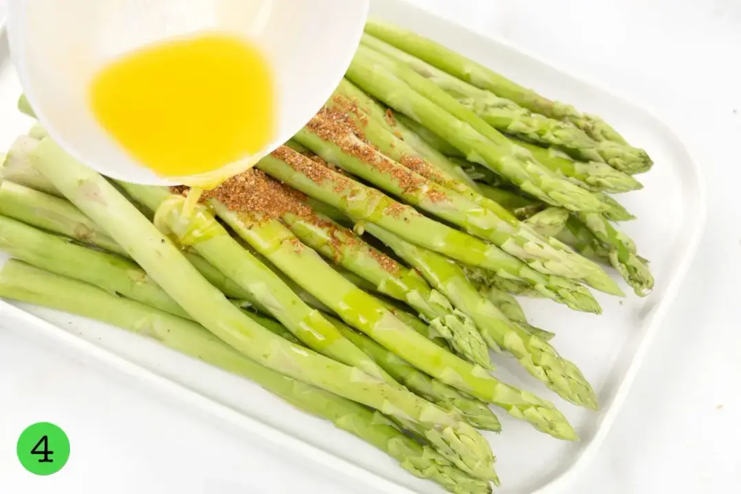 Asparagus stalks topped with a spice blend laying on a plate while melted butter are being poured over them