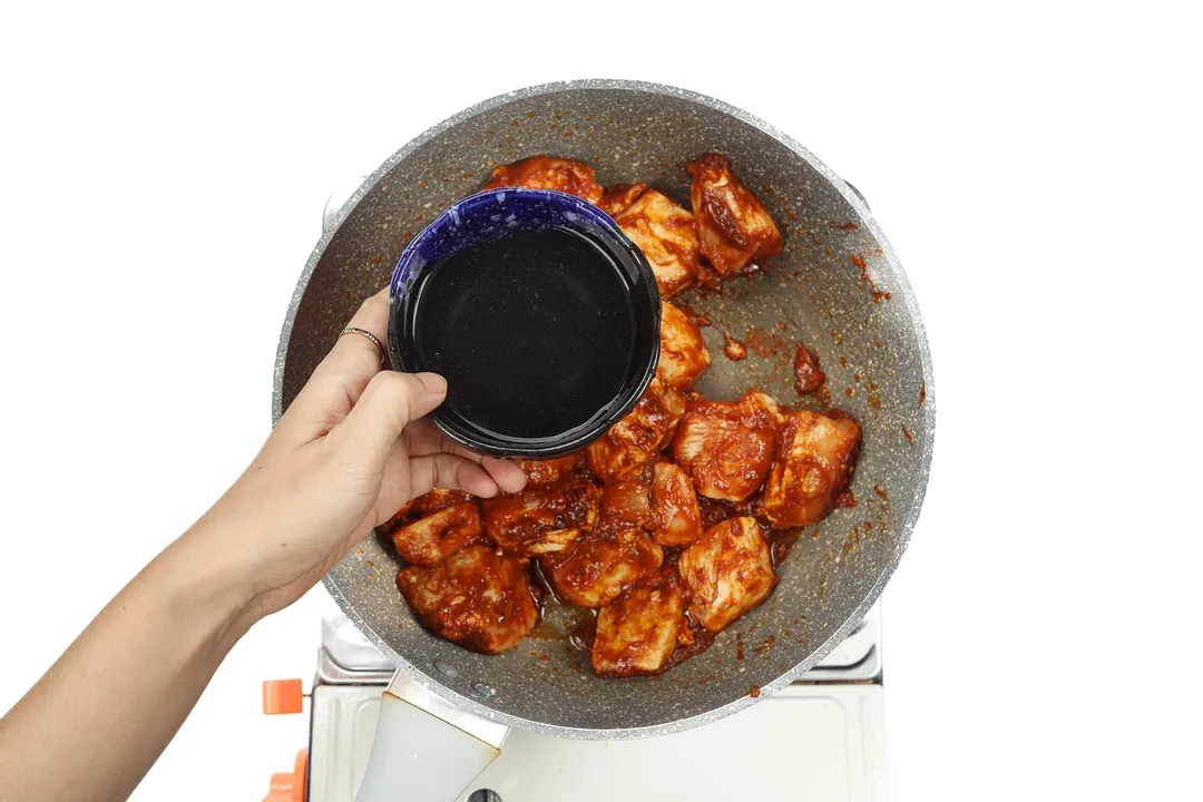A hand holding a blue bowl containing a dark sauce over a large pan cooking chicken cubes covered in spices