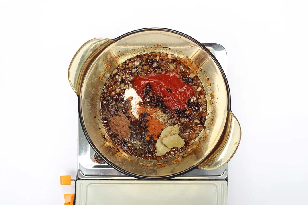 A saucepan containing a dark brown sauce mixture with several spices and red ketchup on top