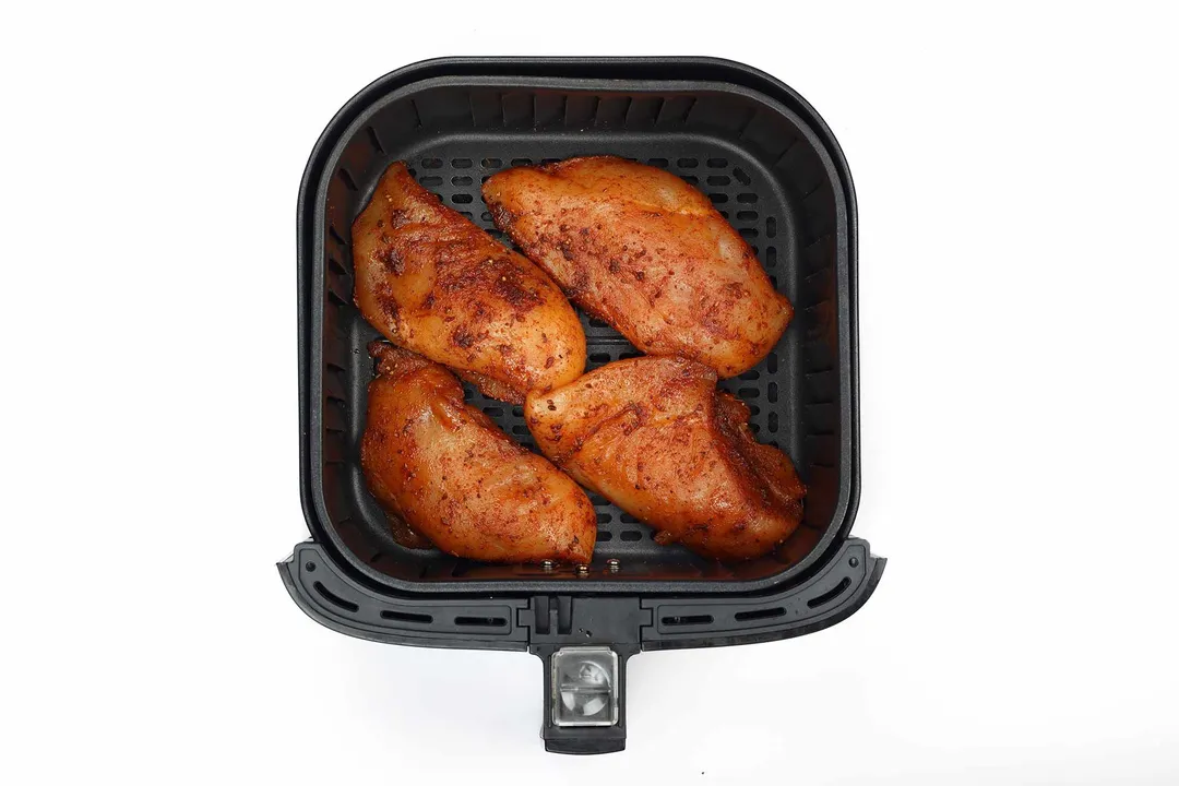 An air fryer basket containing four pieces of raw chicken breast covered in spices