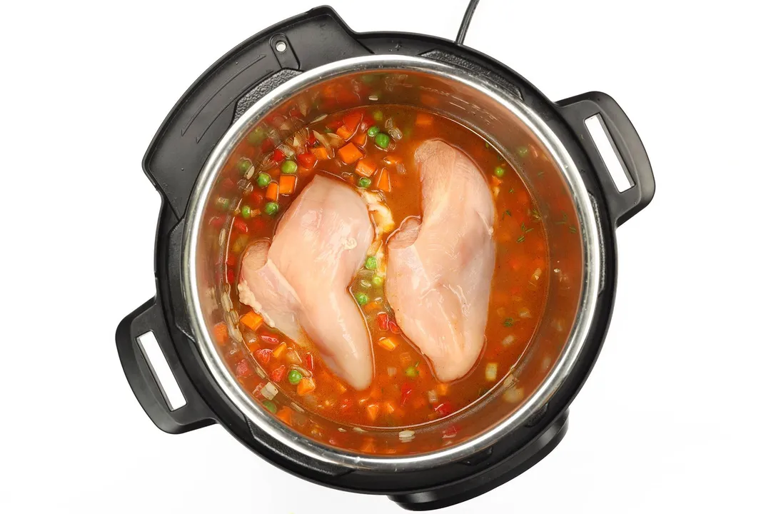 An instant pot's inner pot cooking two raw chicken breasts in a soup with diced vegetables and green peas