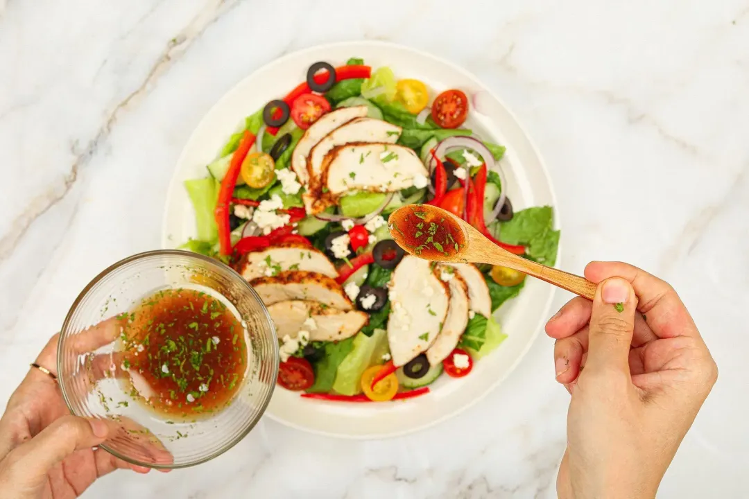 A hand using a wooden spoon to distribute a dark brown dressing all over a Greek Chicken Salad