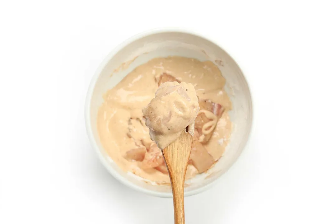 A white bowl holding up a thick and beige batter with a bowl of the same batter and filled with raw chicken cubes in the background