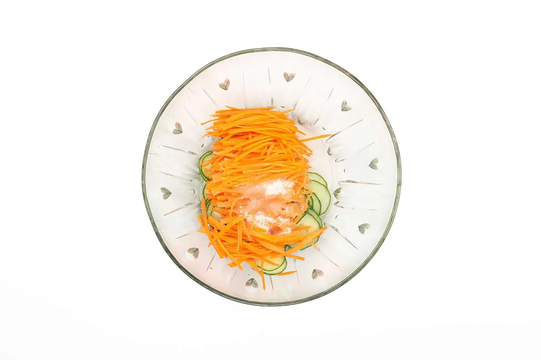 Julienned carrots and sliced cucumbers with pickling ingredients in a bowl