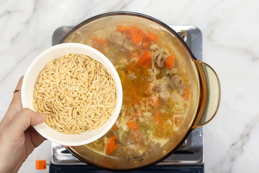 hand holds a bowl of orzo pasta on top of a pot of soup