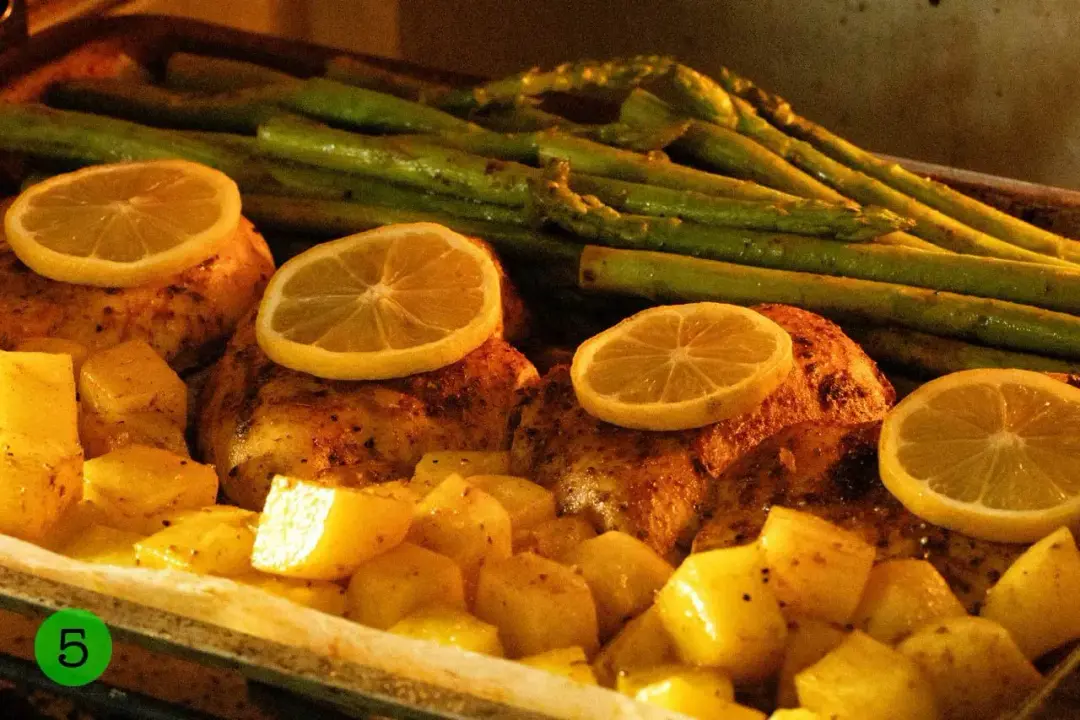 A baking pan filled with chicken breasts, lemon slices, potato cubes, and asparagus stalks being baked in the oven