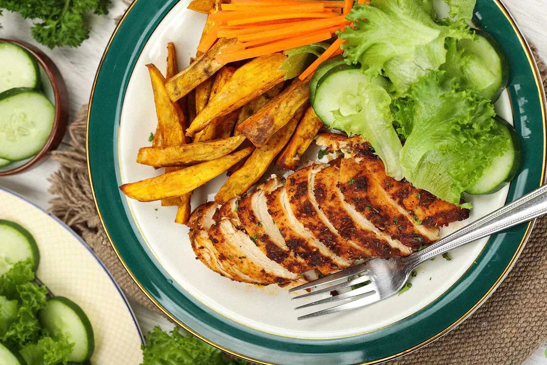A plate containing a crosswise sliced chicken breast, sweet potato fries, julienned carrots, and cucumber slices