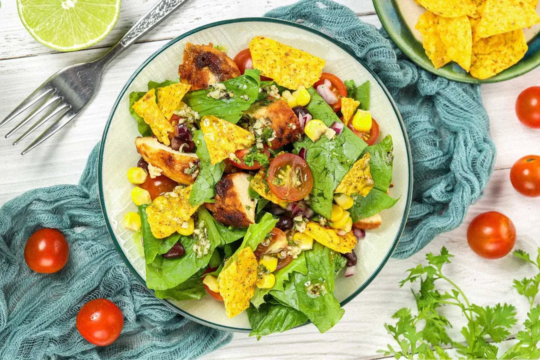 A colorful plate of chicken taco salad filled with dorito chips, lettuce pieces, halved cherry tomatoes, and corn laid on a blue mesh cloth and surrounded by cherry tomatoes