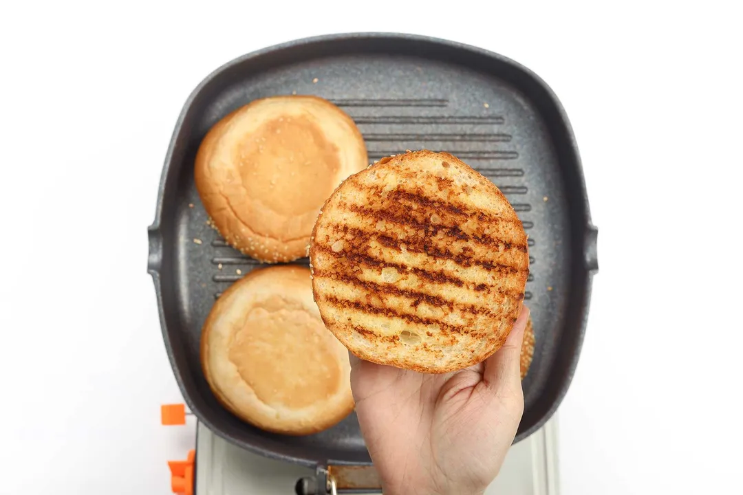 A hand holding a charred burger bun over a griddle that's cooking two hamburger buns