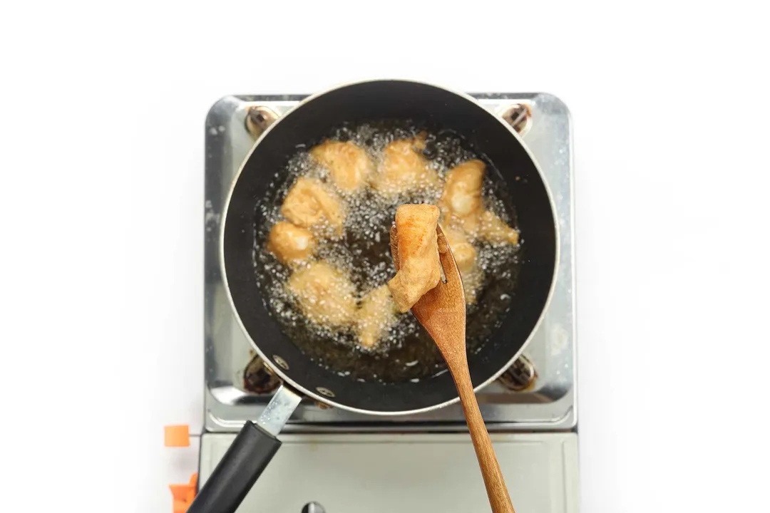 A pan deep-frying chicken cubes and a wooden fork holding a piece of the deep-fried chicken cube up