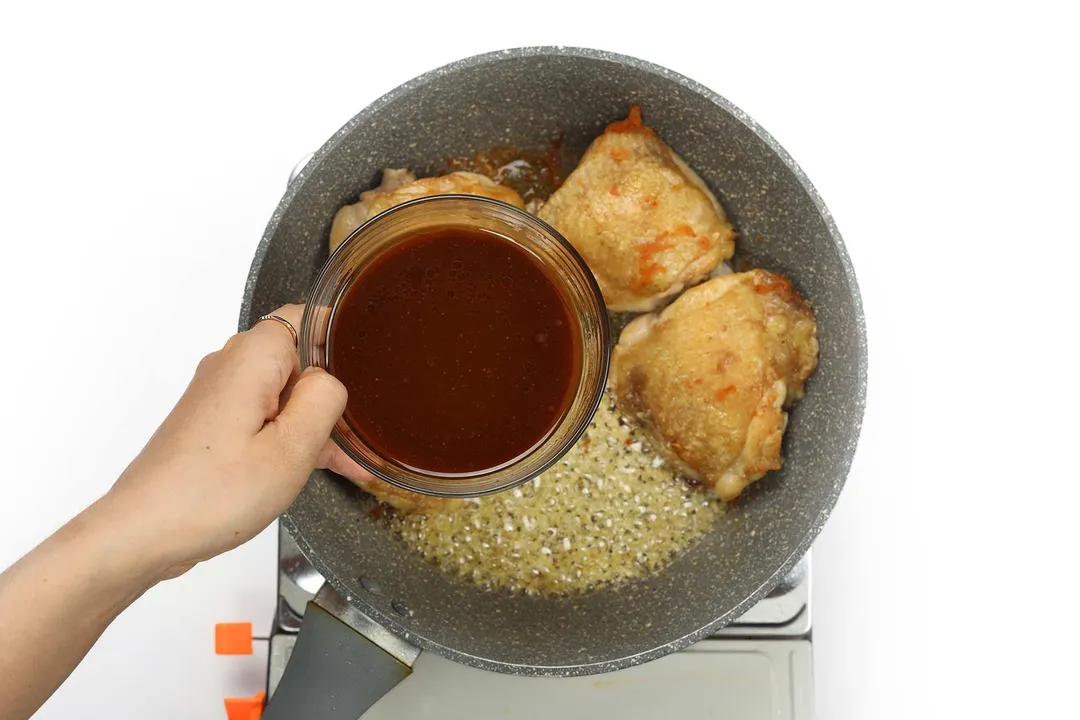 A hand holding a small bowl of miso sauce over a large pan cooking chicken thighs and aromatics