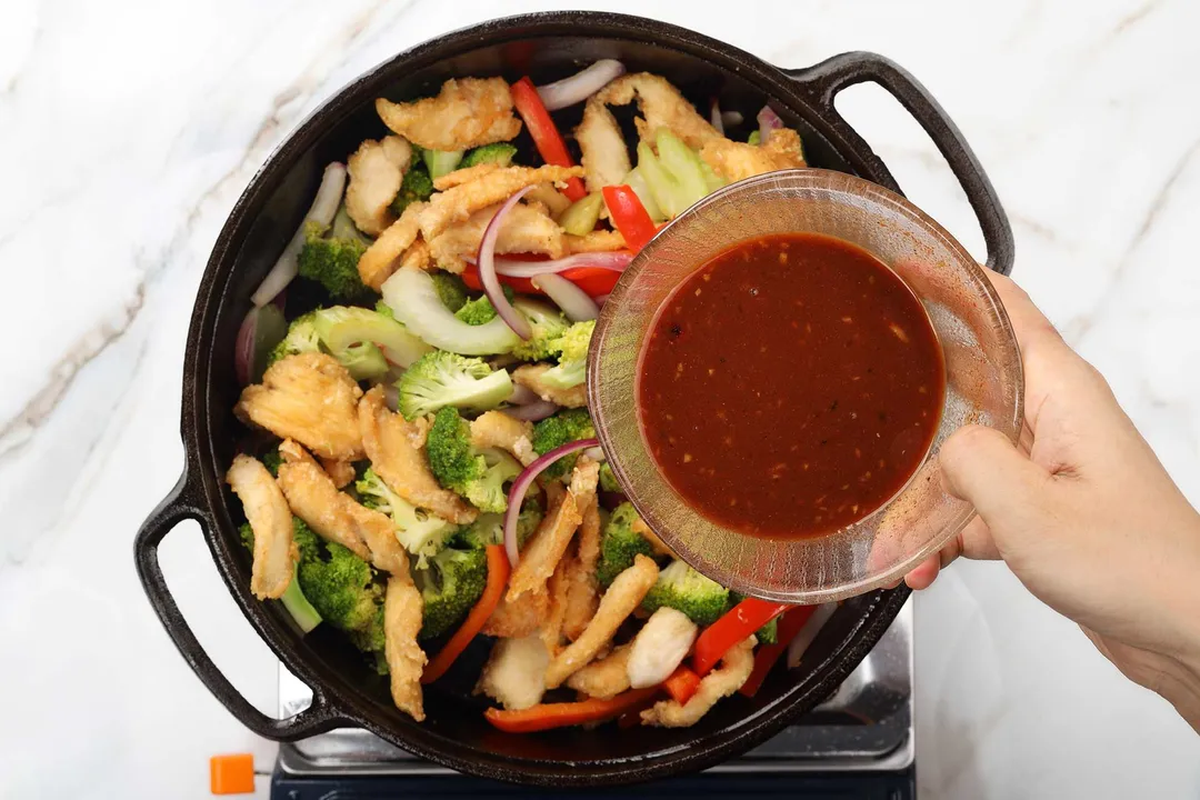 a hand holds a bowl of sauce on top of a cast iron skillet of cooked chicken with vegetables
