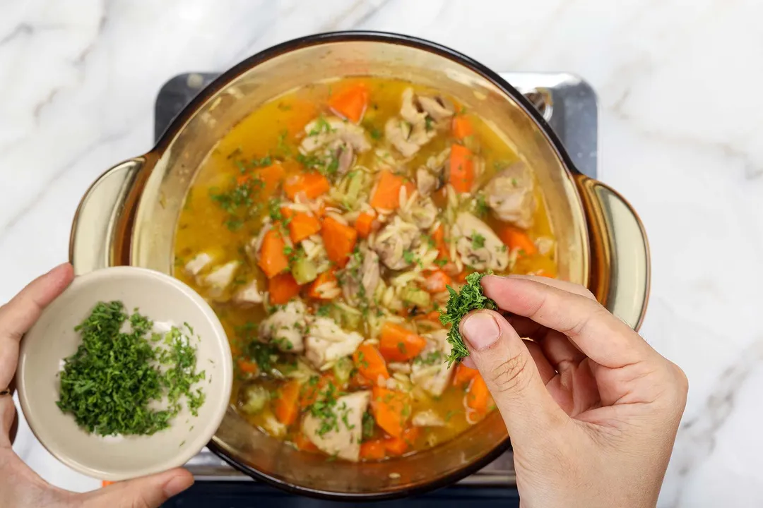 sprinkling chopped parsley from a small bowl onto a pot of chicken orzo soup