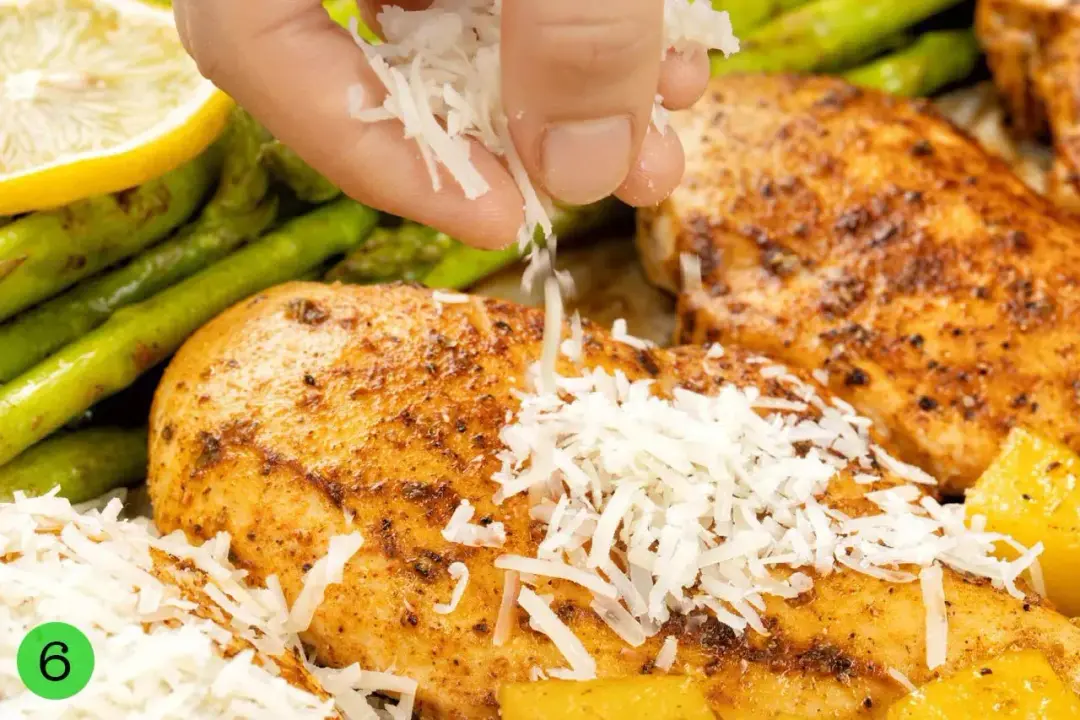 Parmesan cheese being sprinkled over baked chicken breasts that are surrounded by asparagus stalks and lemon slices