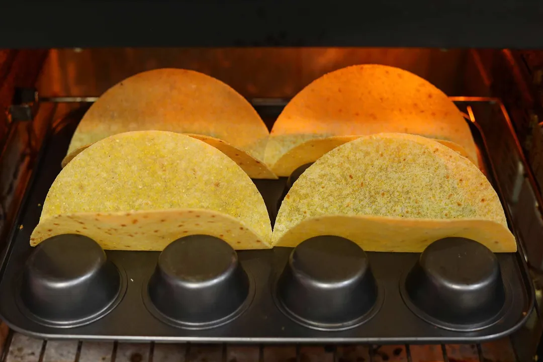 Tortillas folded in the empty spaces of an upside-down muffin tray. The tray is laid in the middle rack of a baking oven