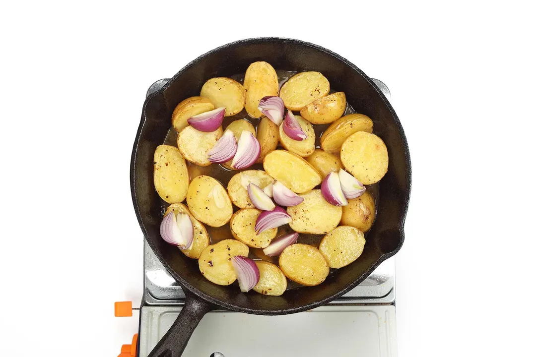 A large black skillet cooking baby potato halves and quartered shallots