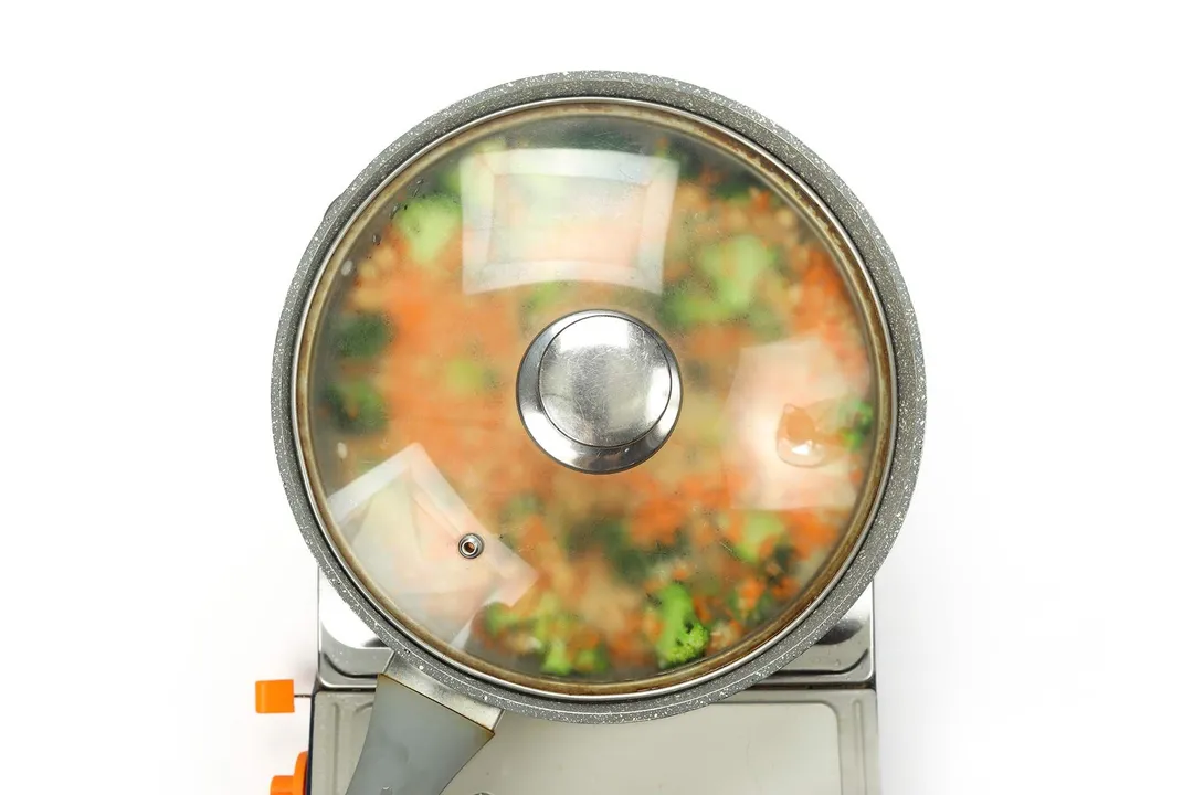 A large pan covered with a glass lid cooking broccoli, carrots, and couscous