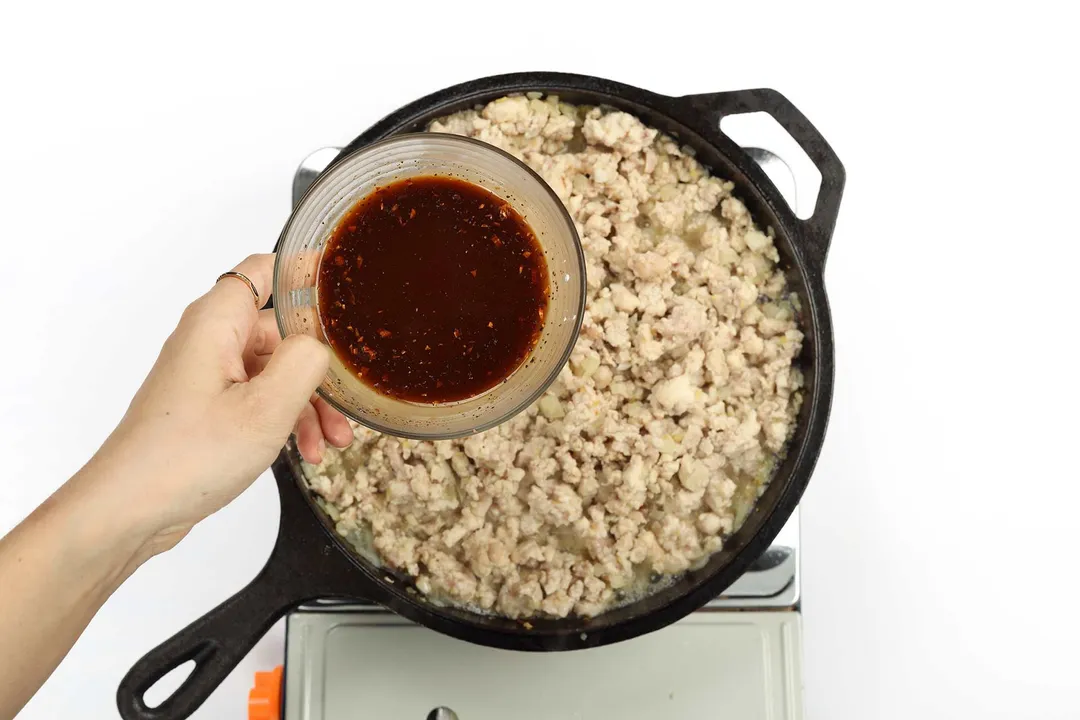 A hand holding a bowl containing a dark red sauce over a skillet filled with cooked ground meat