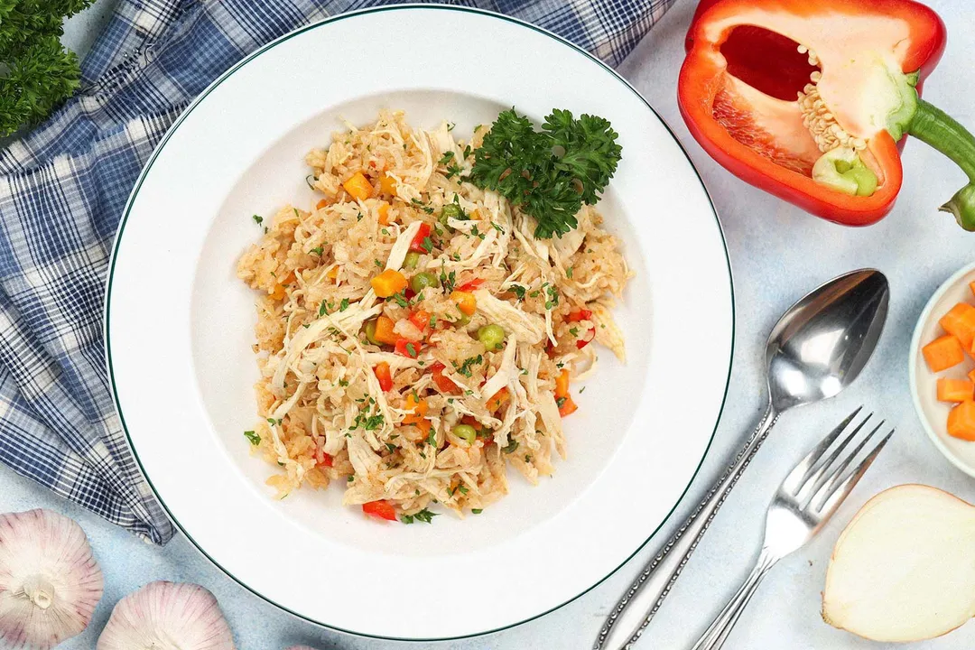 A flatlay of a dish of rice and shredded chicken, a spoon, a fork, half a bell pepper, and garlic