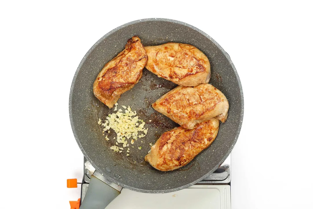 A large pan cooking four pieces of chicken breasts and a pile of minced garlic