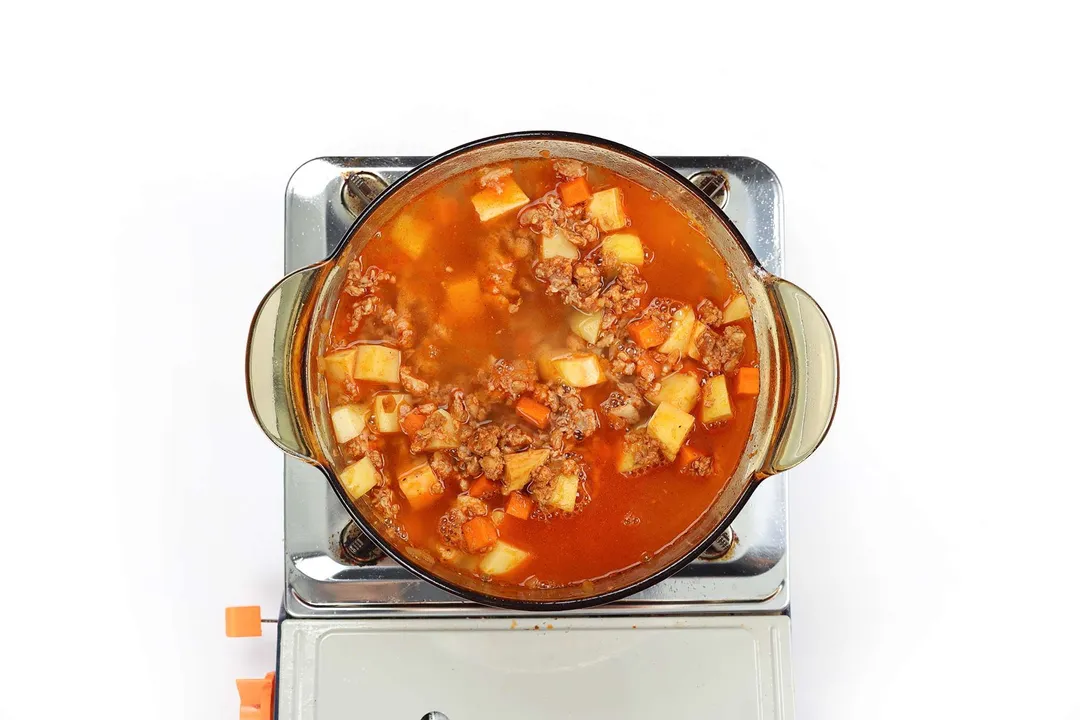 A glass saucepan cooking a ground chicken soup filled with chicken broth, cubed potatoes, coarsely diced carrots, and ground chicken