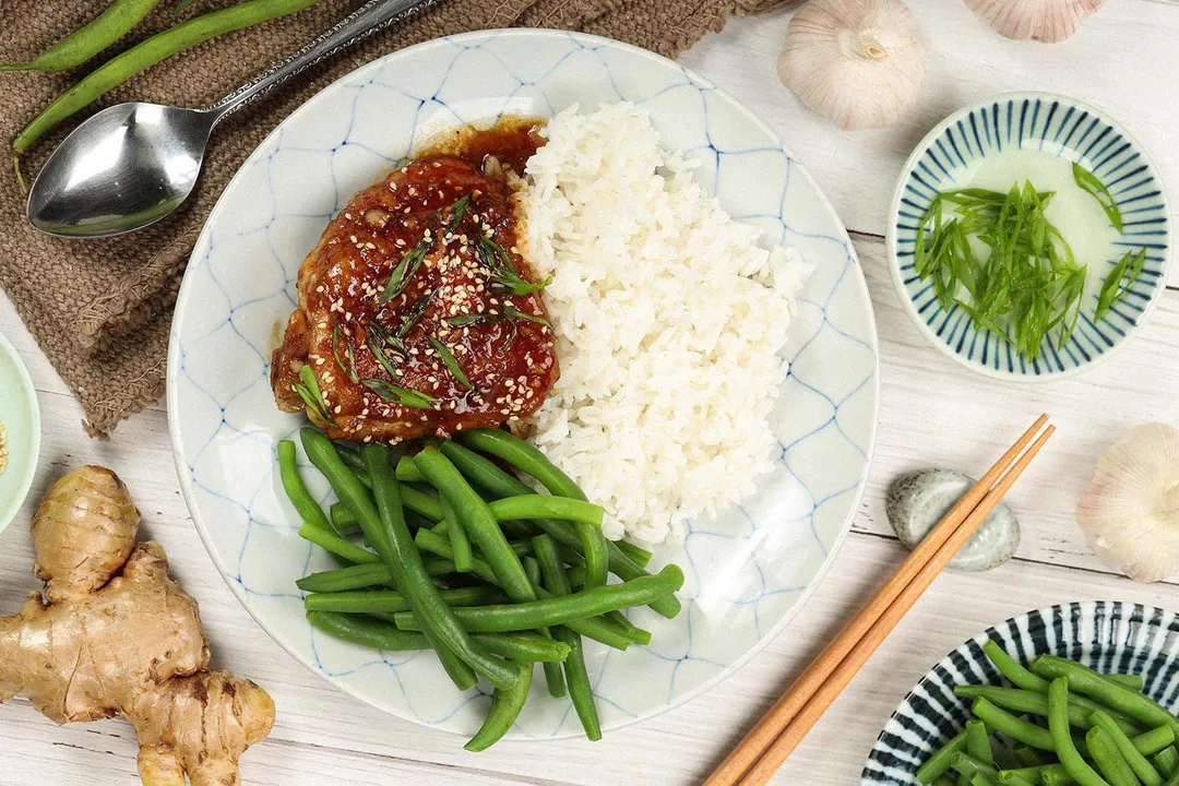 A high angle shot of a plate containing miso chicken thigh, white rice, and green beans laid near garlic cloves, a ginger, a spoon, a brown tablecloth, and a small dish of chopped scallions