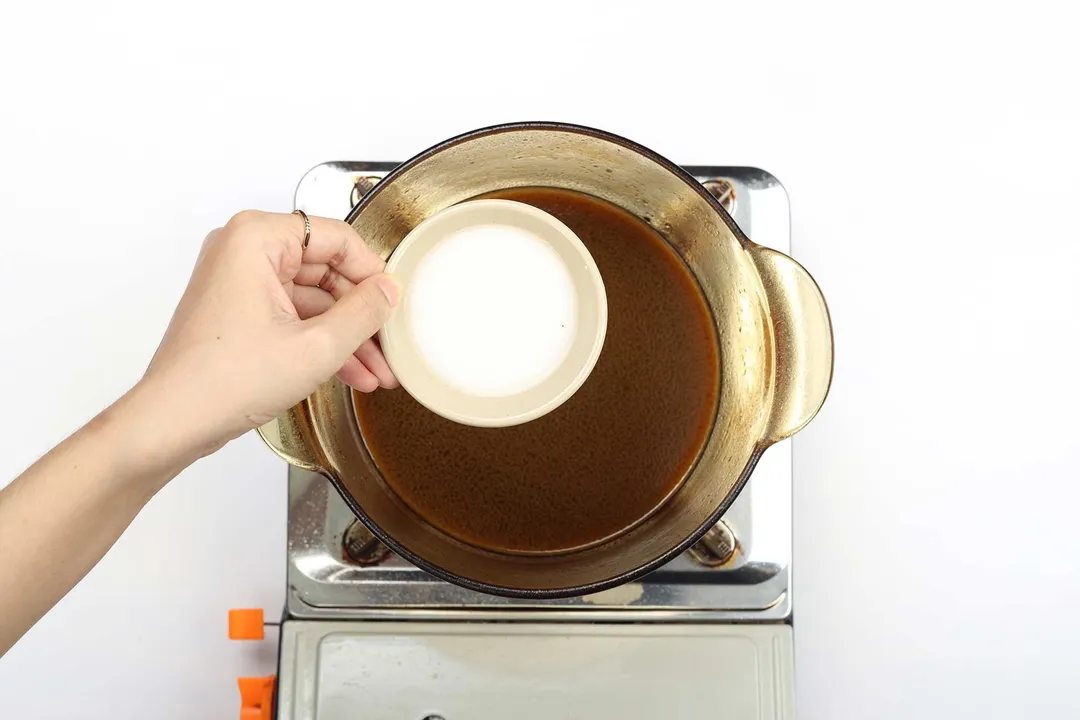 A hand holding a sauce dish containing white flour over a saucepan that's cooking a dark brown sauce