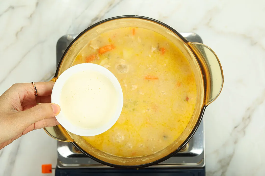 hand holds a small bowl of heavy cream on top of a pot of soup