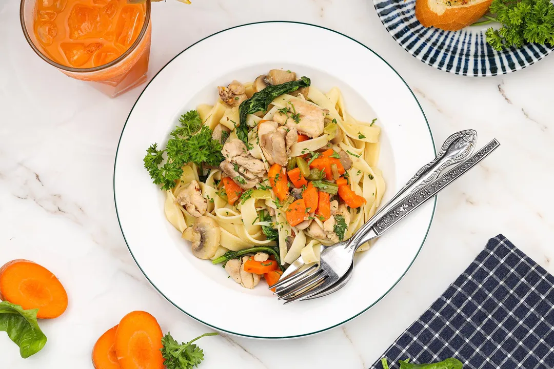 a plate of cooked chicken noodle soup next to a glass of carrot juice a bread