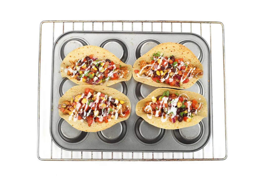 Four tacos laid in the empty spaces of an upside-down muffin tray. The tray is laid on top of another baking rack.