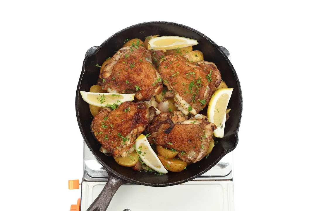 A large black skillet cooking four chicken thighs with browned skin and four lemon wedges