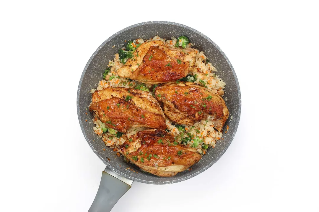 A gray skillet of golden brown chicken thighs sitting atop cooked broccoli couscous