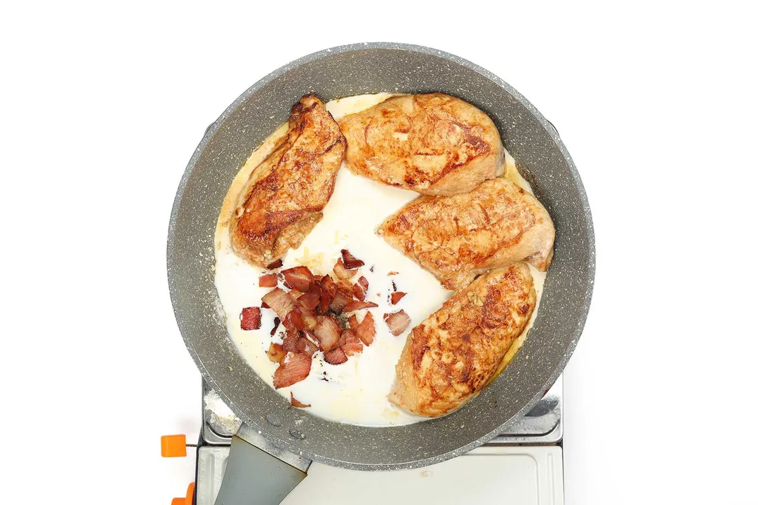 A large pan containing four pieces of cooked chicken breasts and a pile of bacon drenched in a white cream sauce