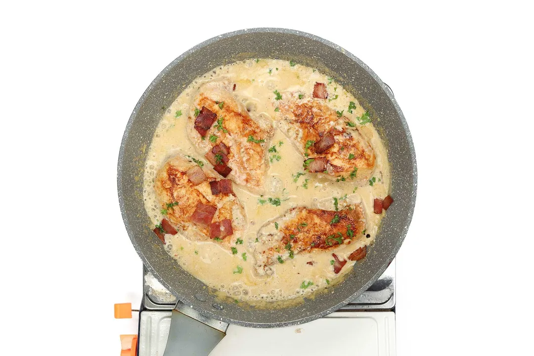 A large pan cooking four pieces of cooked chicken and bacon pieces drenched in a pale yellow cream sauce