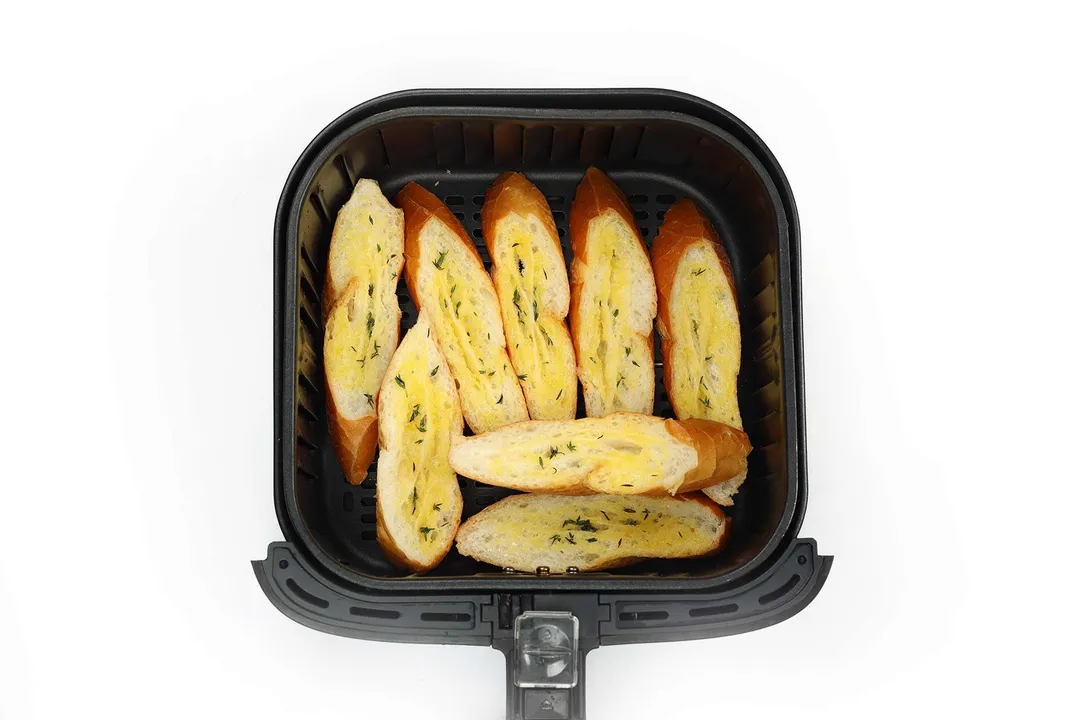 An air fryer basket containing slices of baguette swept with butter and chopped herbs