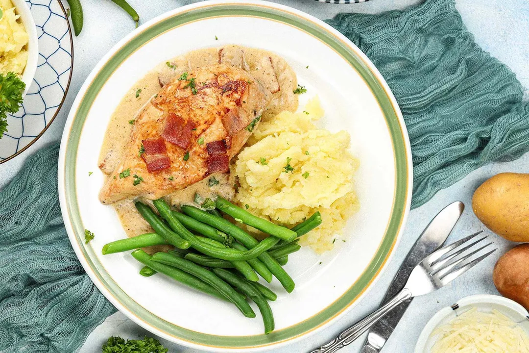 A plate containing a piece of chicken breast, mashed potatoes, and green beans placed on top of a teal mesh cloth and next to a fork