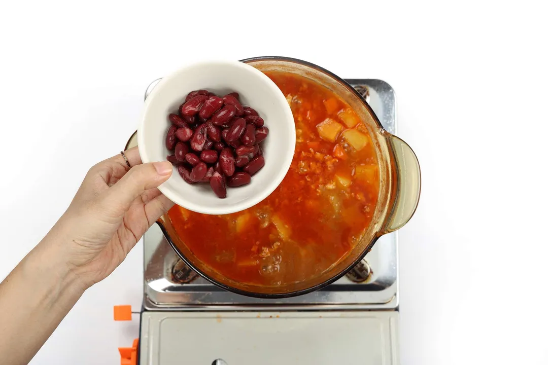 A hand holding a small white bowl containing kidney beans to pour into a glass saucepan that contains ground chicken soup