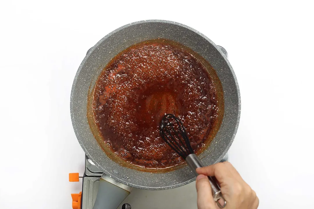 A hand whisking a dark, thick, and glossy honey sauce that’s bubbling in a pan 