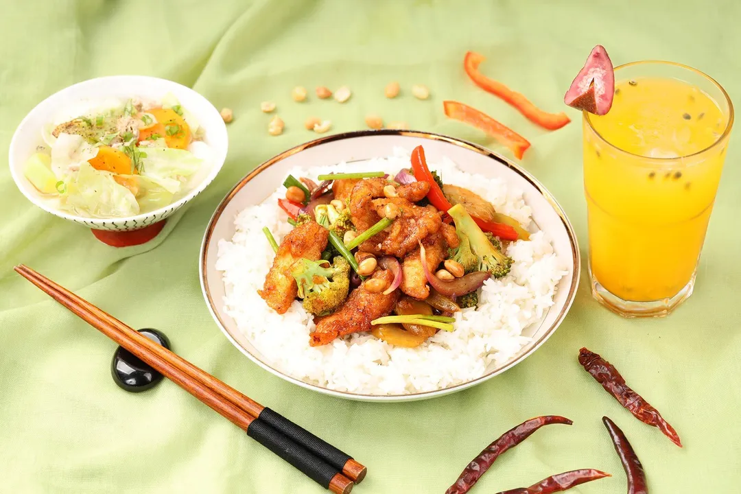 a plate of cooked hunan chicken and rice next to a glass of juice a a bowl of vegetable soup