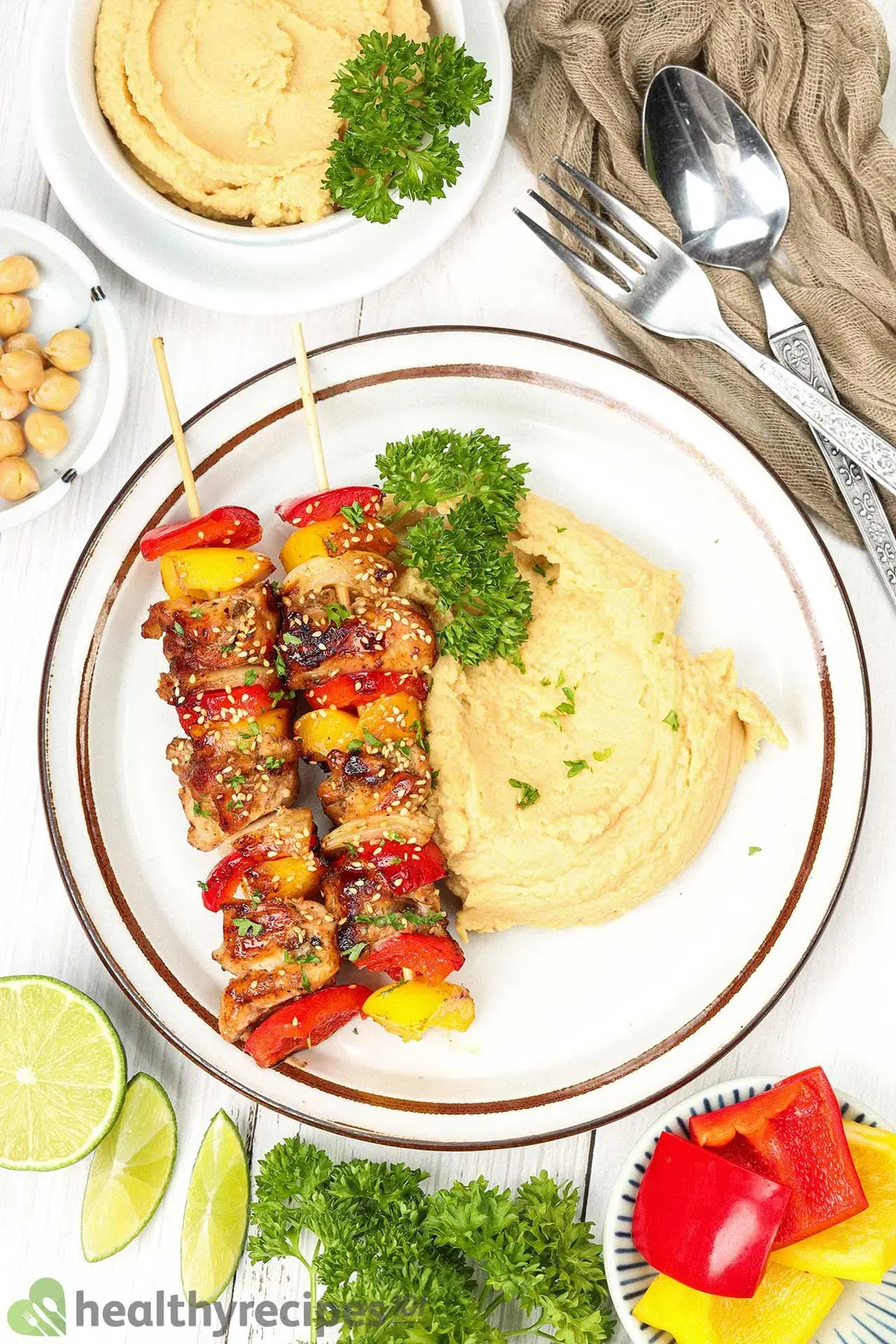 A top-down view of a plate of chicken skewers and chickpea puree alongside a bowl of chickpea puree and other utensils