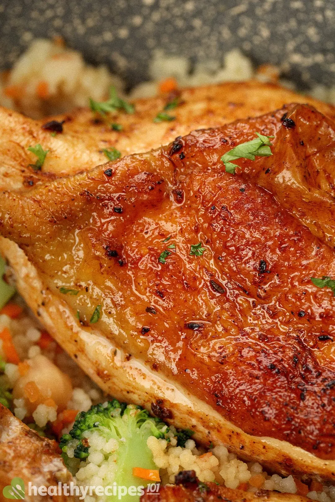 A close-up shot of a cooked chicken thigh whose skin is deliciously browned with a few charred marks, laid on a bed of couscous and near a floret