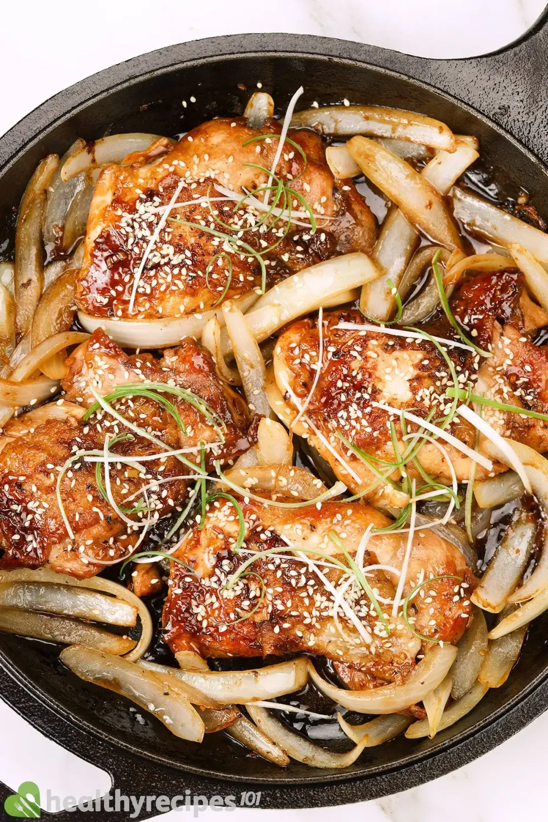 A large cast iron skillet of glossy chicken thighs with white sesame seeds, sliced white onions, and shaved green onions