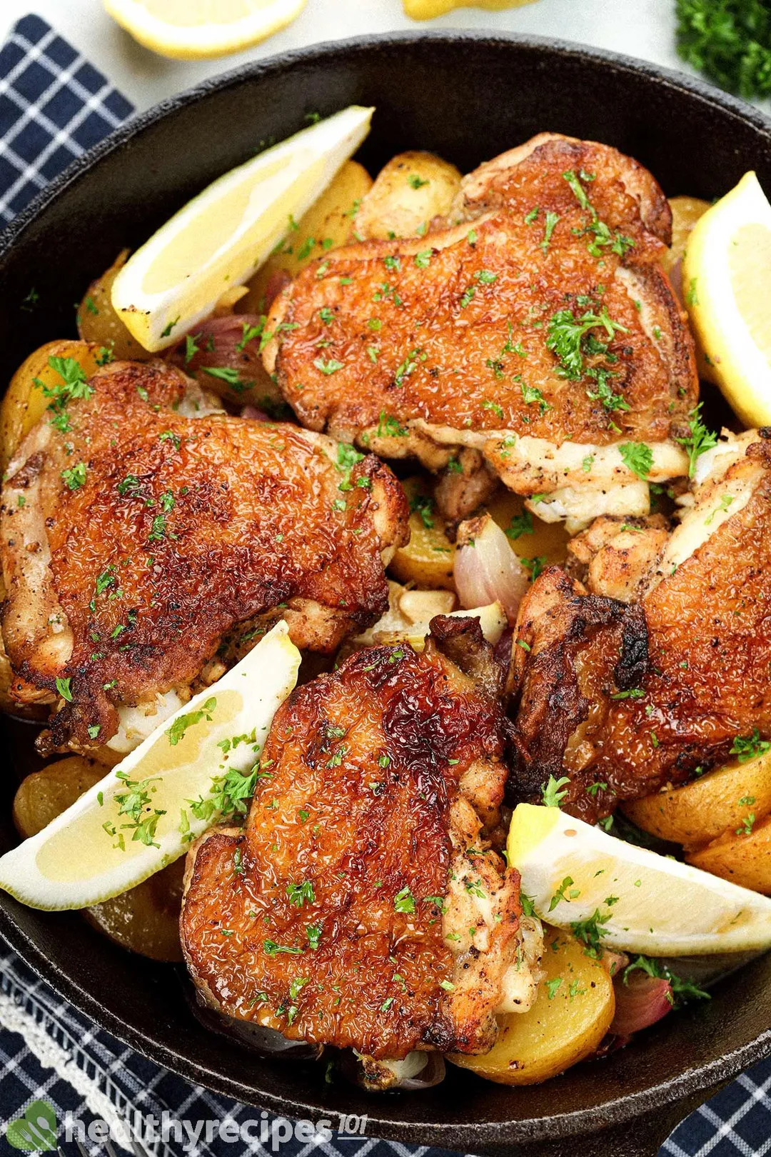 A deep skillet filled with browned and cooked chicken thighs, lemon wedges, and baby potato halves