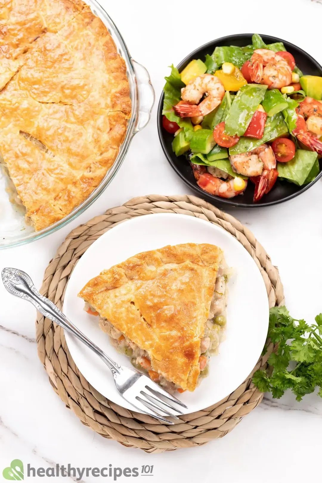 A high angle shot of a slice of chicken pot pie placed near a bowl of salad, a fork, and fresh parsley