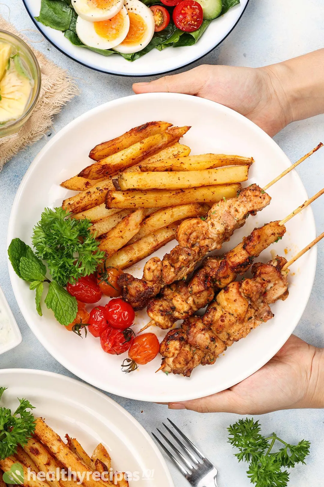 hand holds a plate of cooked chicken skewers and potato batonnet, cherry tomato