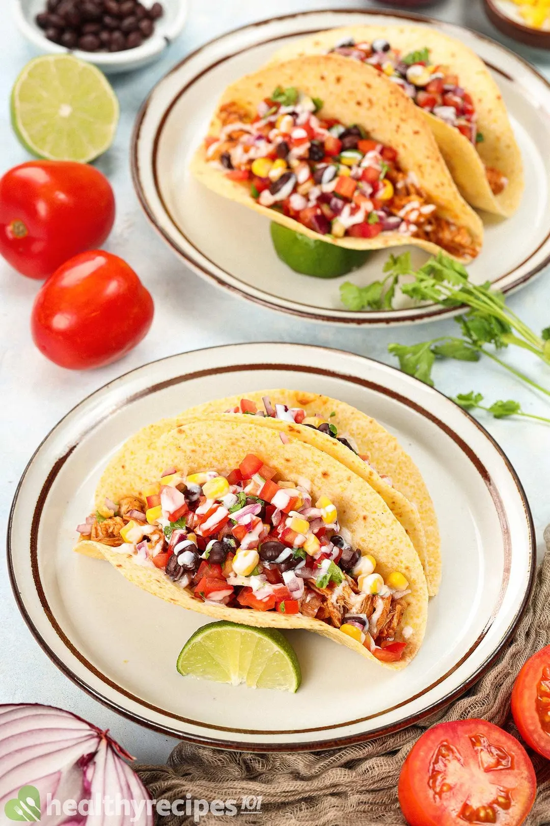 Two plates containing BBQ chicken tacos laid near tomatoes, red onions, fresh coriander, and a brown mesh cloth