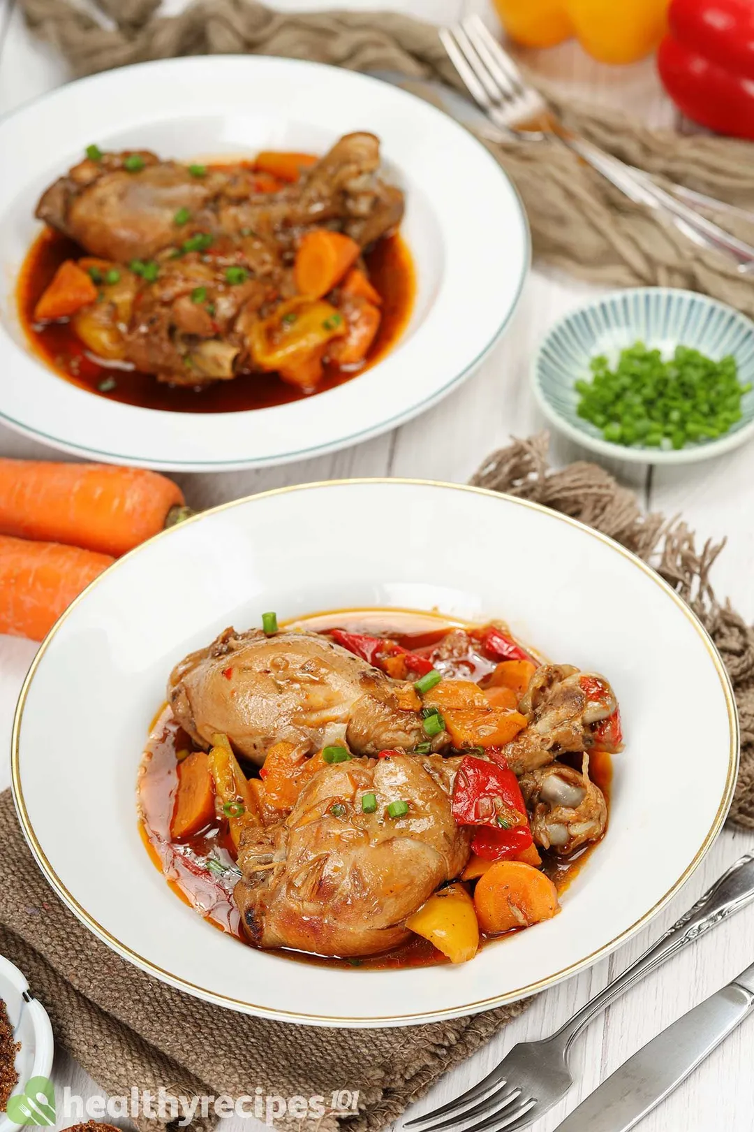 Two plates of brown stew chicken placed on a table decorated with a fork, a dinner knife, and a cloth