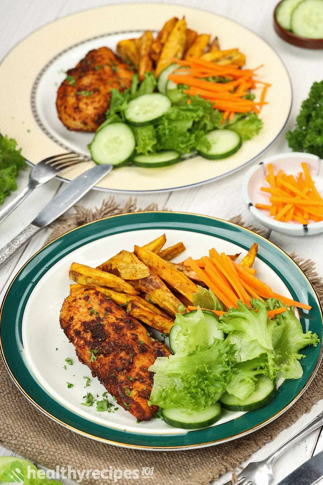 Two plates containing cooked chicken breasts covered in spices, sweet potato fries, cucumber slices, julienned carrots, and lettuce leaves